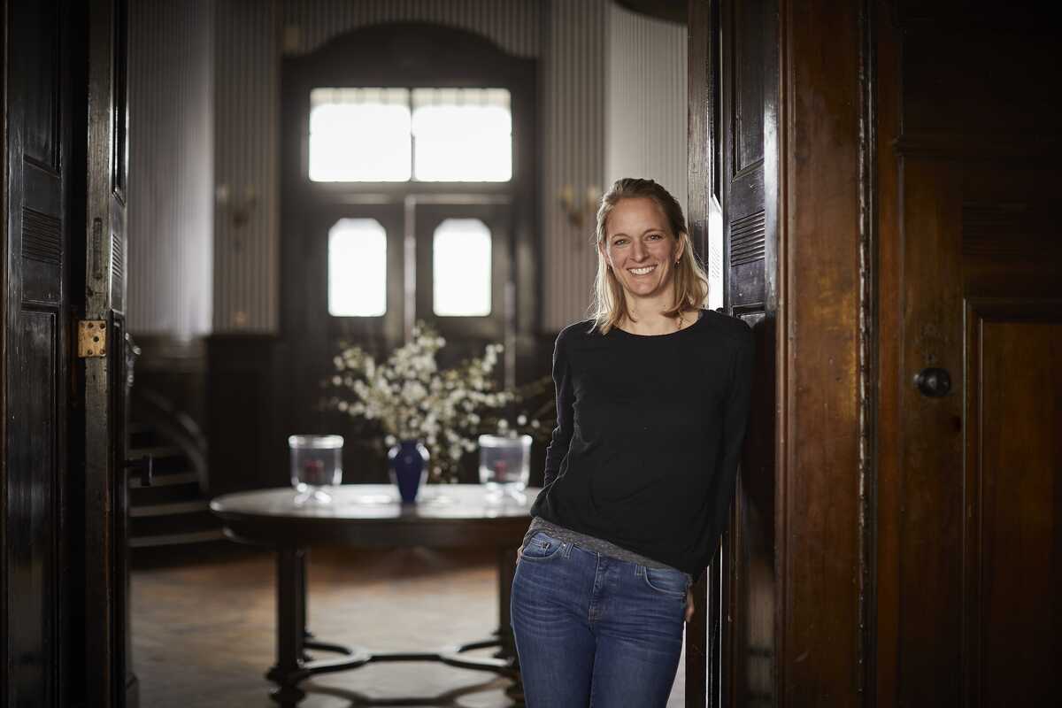 Foto: Marco Warmuth_TGZ/Hayessen - Eventlocation Rittergut Etzdorf Eine Frau steht in einem großen Raum an einer Holztür. Hinter ihr steht ein Tisch mit Blumen. Dahinter ist eine weitere Tür mit Fenstern.